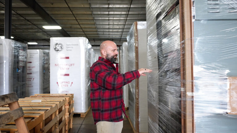 Erik Thompson stands and holds his right hand up in the air pointed toward a shelf filled with switchboards encased in transparent plastic wrapping, with more wrapped stacks of packaged items in rows behind him and a row of wood crates, in a room with a metal ceiling. Erik Thompson stands and holds his right hand up in the air pointed toward a shelf filled with switchboards encased in transparent plastic wrapping, with more wrapped stacks of packaged items in rows behind him and a row of wood crates, in a room with a metal ceiling.
