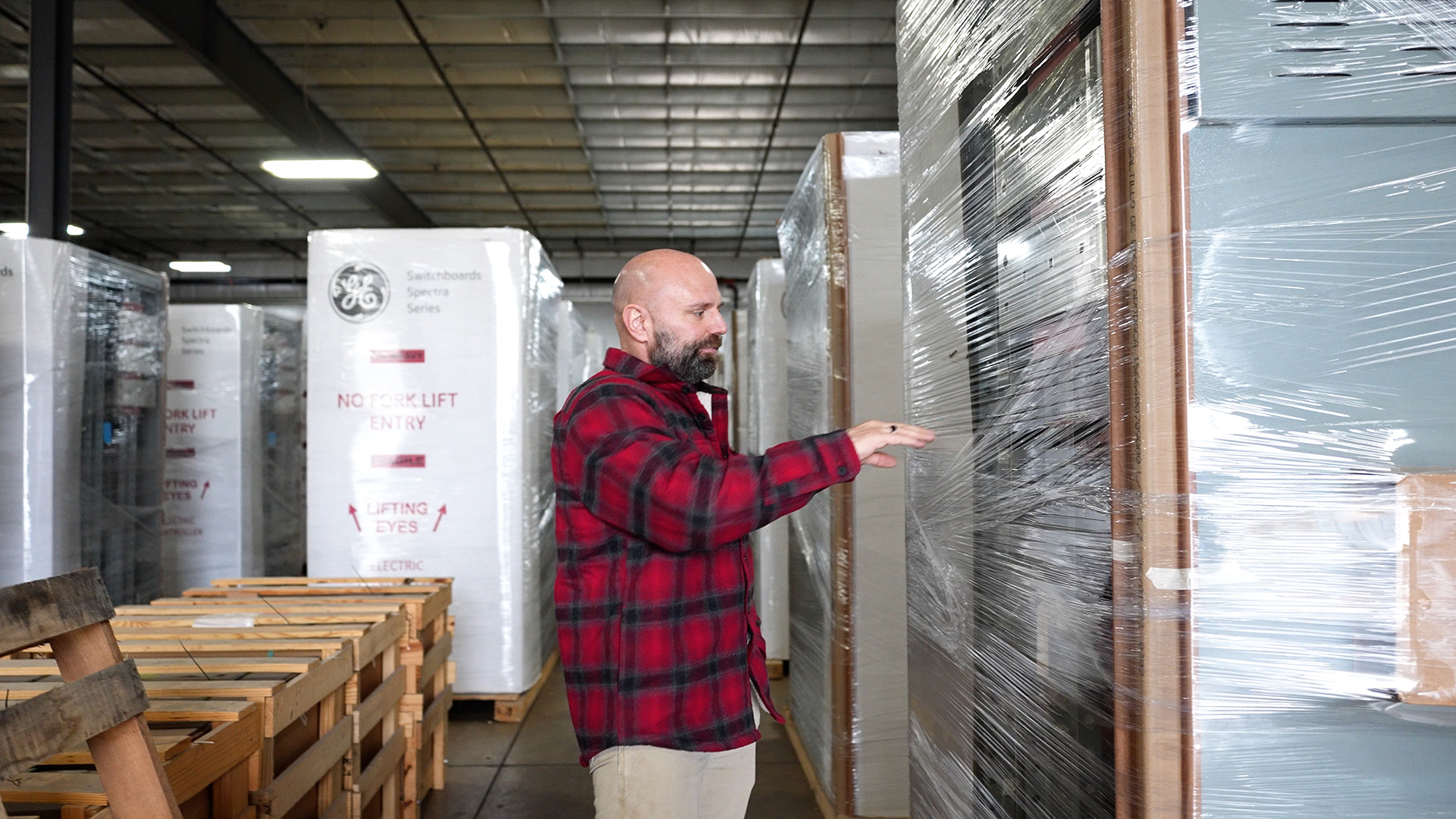 Erik Thompson stands and holds his right hand up in the air pointed toward a shelf filled with switchboards encased in transparent plastic wrapping, with more wrapped stacks of packaged items in rows behind him and a row of wood crates, in a room with a metal ceiling.