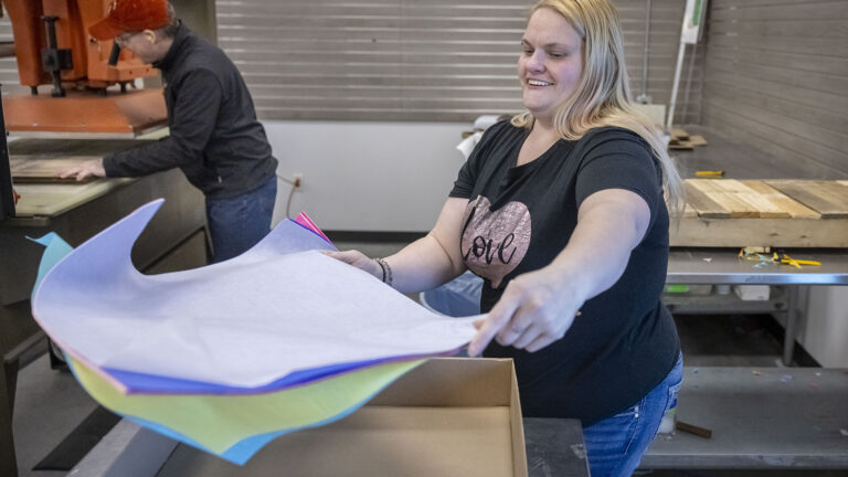 Mellisa Edwards holds multiple sheets of different colors of tissue paper in both hands over a cardboard box while standing behind a metal bench, with a wood pallet on the top of a metal bench behind her and another person standing next to a piece of industrial equipment in the background, in a room with metal shutters along two walls. Mellisa Edwards holds multiple sheets of different colors of tissue paper in both hands over a cardboard box while standing behind a metal bench, with a wood pallet on the top of a metal bench behind her and another person standing next to a piece of industrial equipment in the background, in a room with metal shutters along two walls.