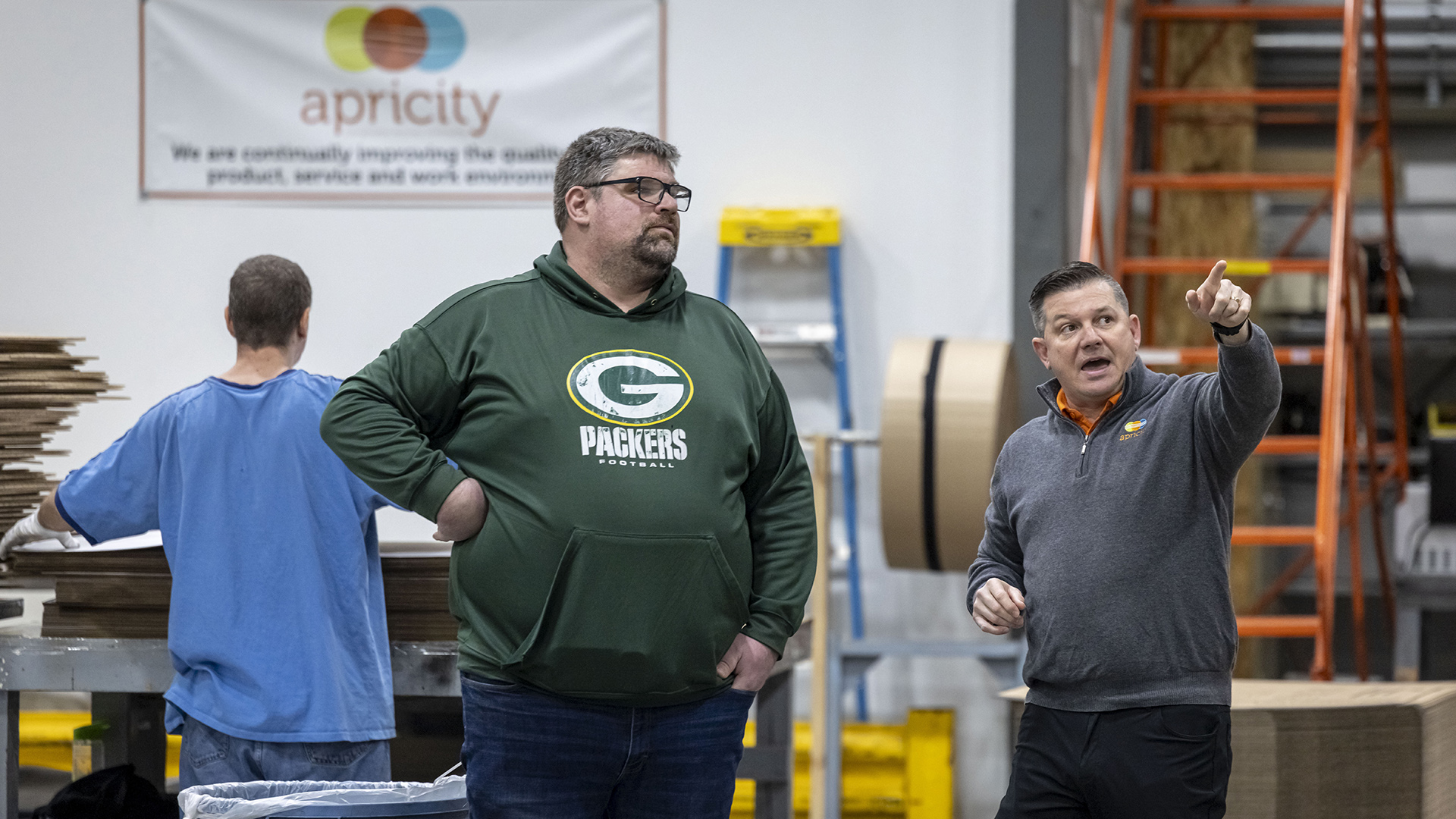 Andy Geurden stands next to Dan Haak, who is pointing with the index finger of his left hand in the direction he is facing, with another person standing behind them facing a stack of flat packaging products on a work table, in a room with stacks and roles of other packaging products next to multiple ladders and a wall-mounted vinyl sign with the word "apricity."