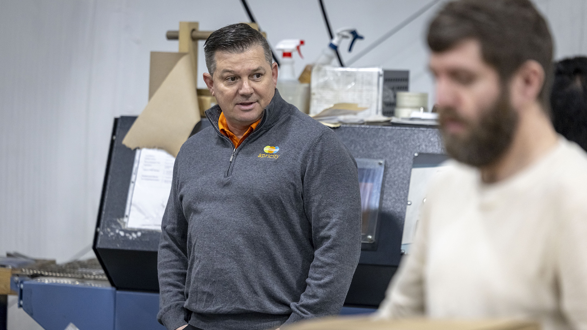 Dan Haak stands with his hands in his pockets, with an out-of-focus person in the foreground and industrial equipment with paper towels, spray bottles and other items on its surface.