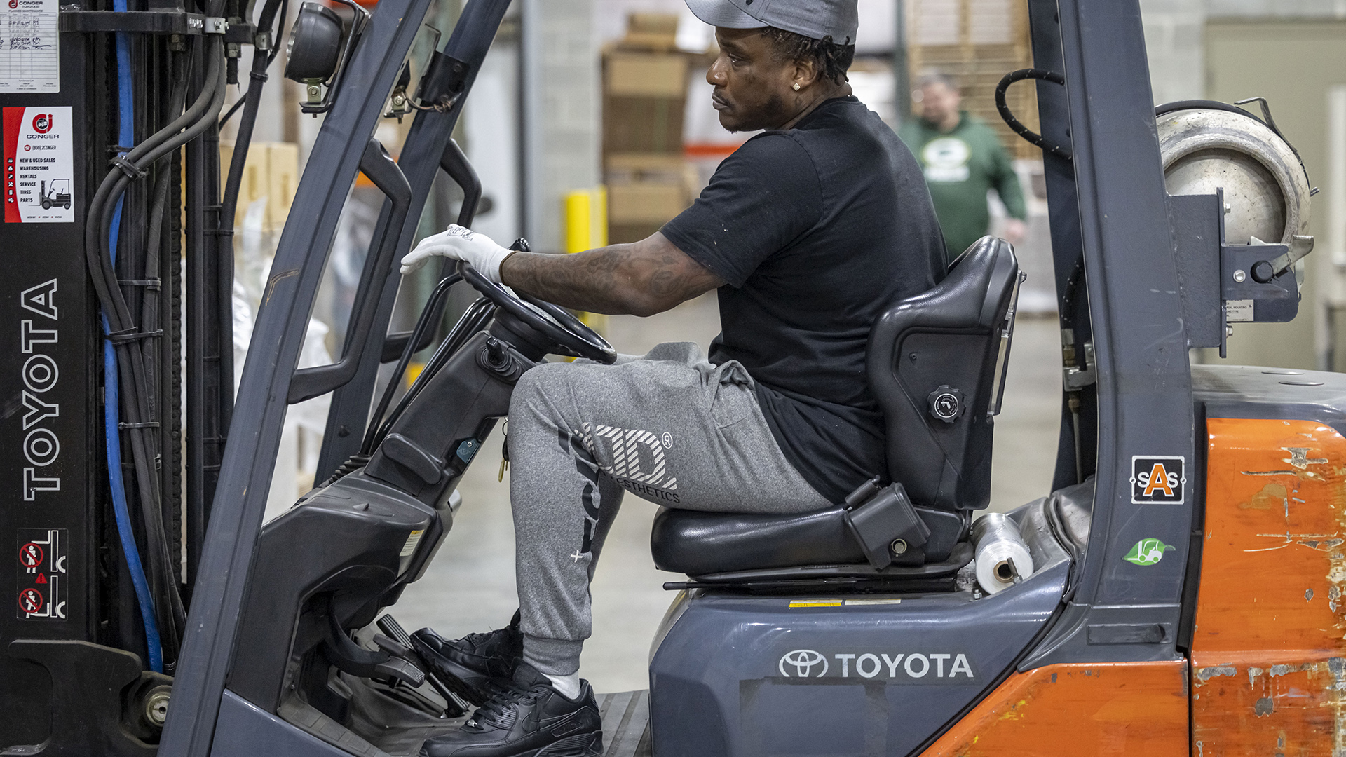 Lindey Jackson sits in the driver's seat in an open-sided forklift with multiple Toyoto wordmarks on it, with an out-of-focus person standing in the background in a room with a concrete floor.