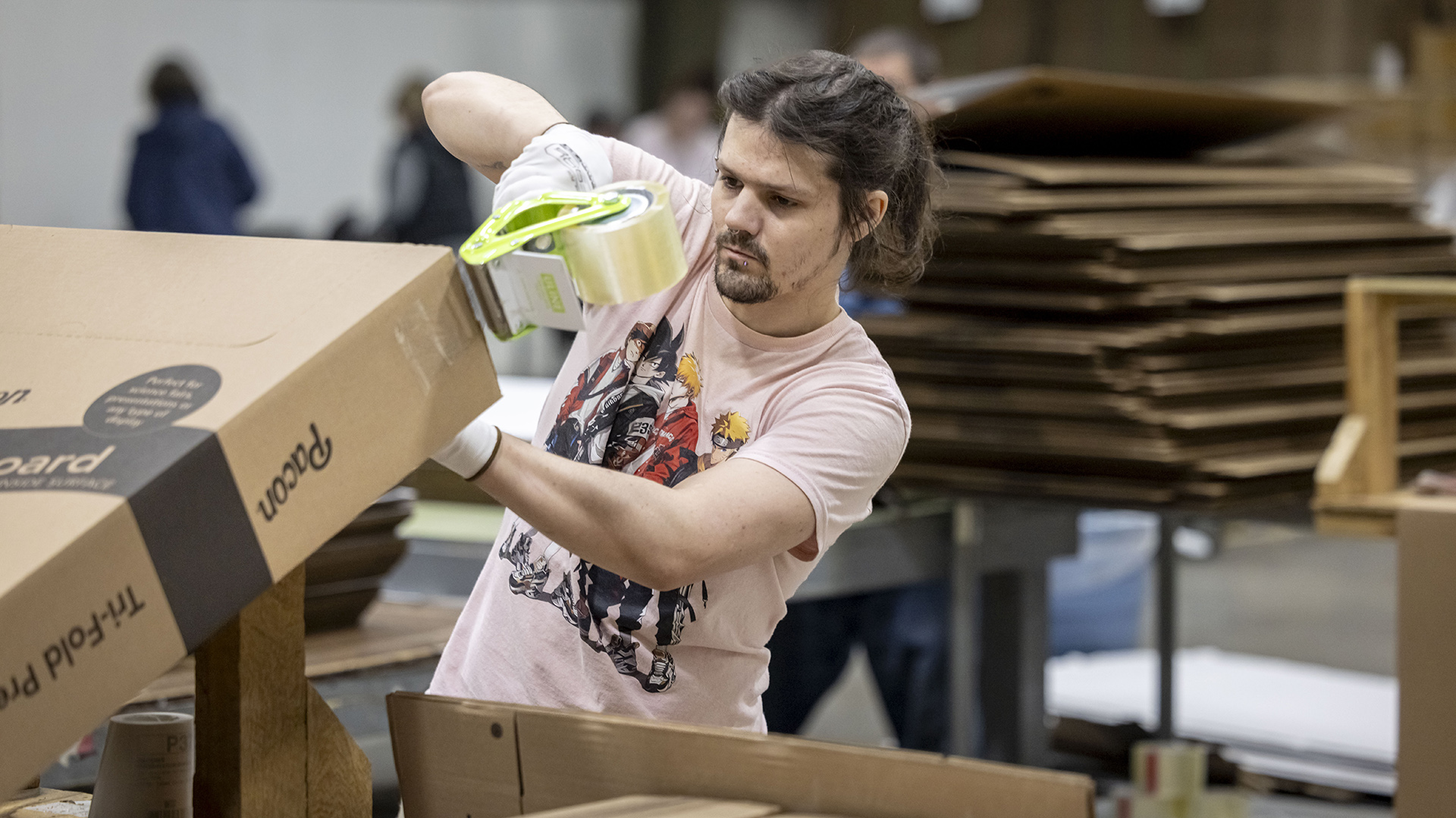Derik Skorbier uses a tape gun dispenser to assemble a cardboard box, with stacks of flat cardboard boxes on a metal bench behind him and out-of-focus people standing in the background.