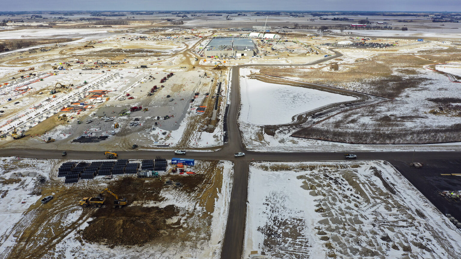 An aerial photo shows a crossroads in the midst of an expanse of partially snow-covered dirt fields with parked vehicles along construction equipment and materials in different locations, with a large building and multiple construction cranes at the far end, with farm fields and stands of trees extending to the horizon.
