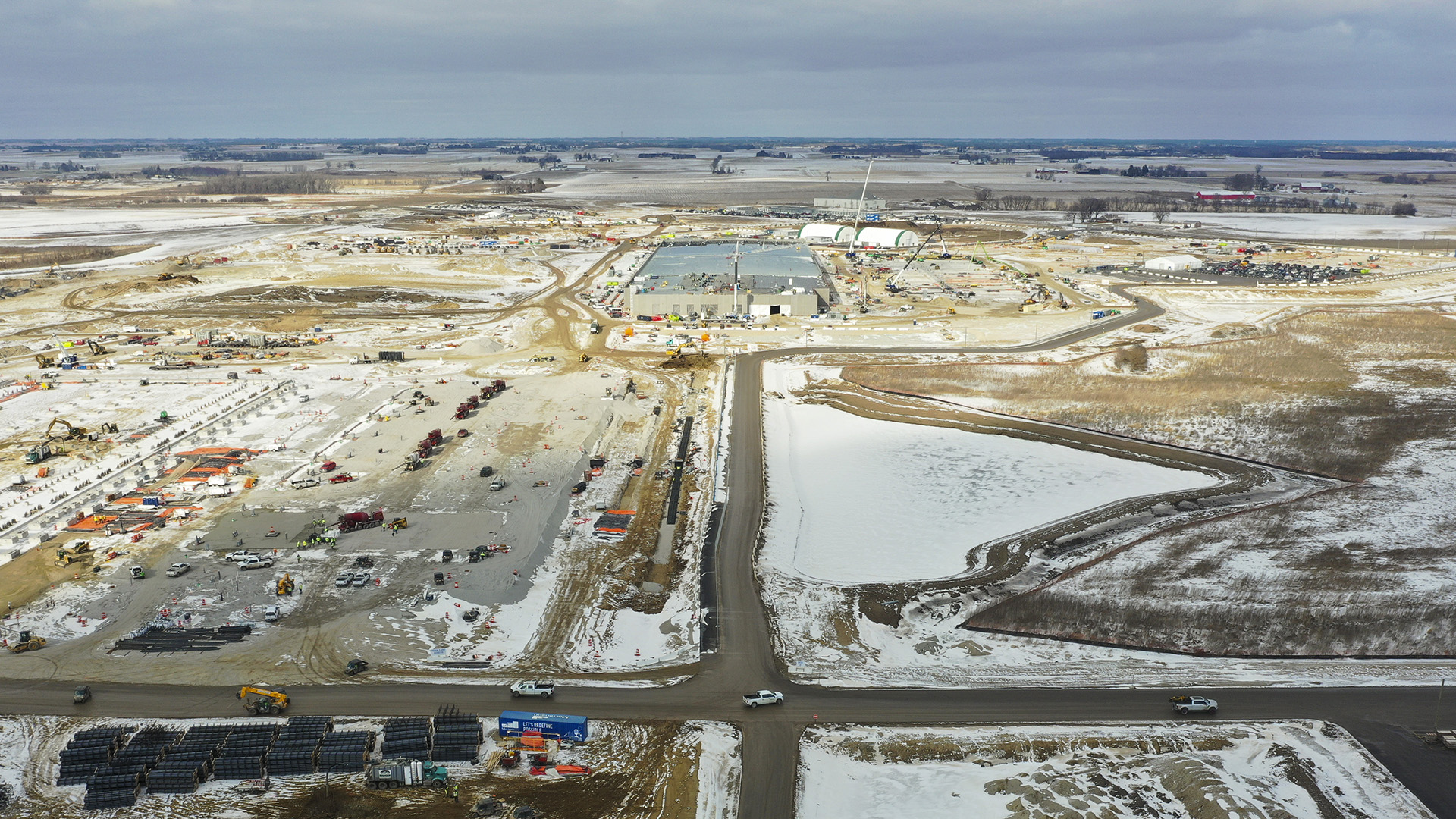 An aerial photo shows a crossroads in the midst of an expanse of partially snow-covered dirt fields with parked vehicles along construction equipment and materials in different locations, with a large building and multiple construction cranes at the far end, with farm fields and stands of trees extending to the horizon.