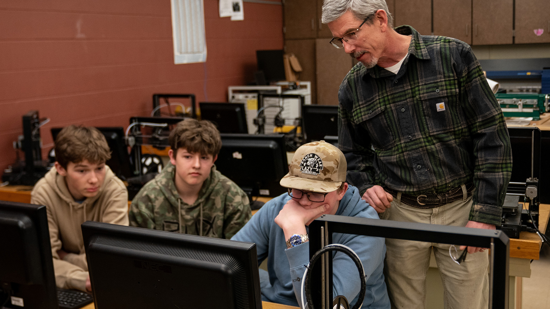Laurence Charlier stands behind three students seated and facing two computer monitors on the surface of a table, with another table behind them with computer monitors and other items on its surface in a room with painted concrete block walls and a row of cabinets on the rear wall.