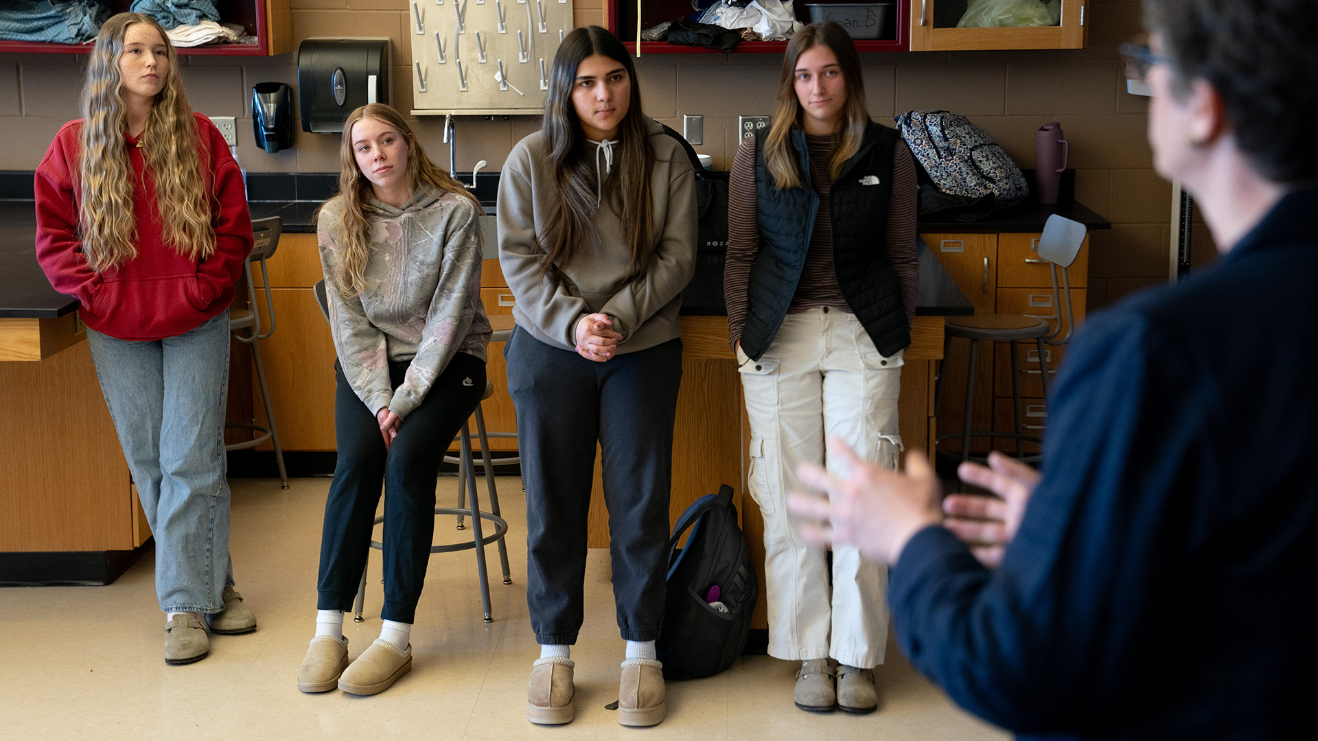 Three students lean against table surfaces and one sits on a round stool while facing Jill Underly, who is out of focus in the foreground, in a room with rows of scientific benches, a sink and drying rack for glassware and wall-mounted cabinets.