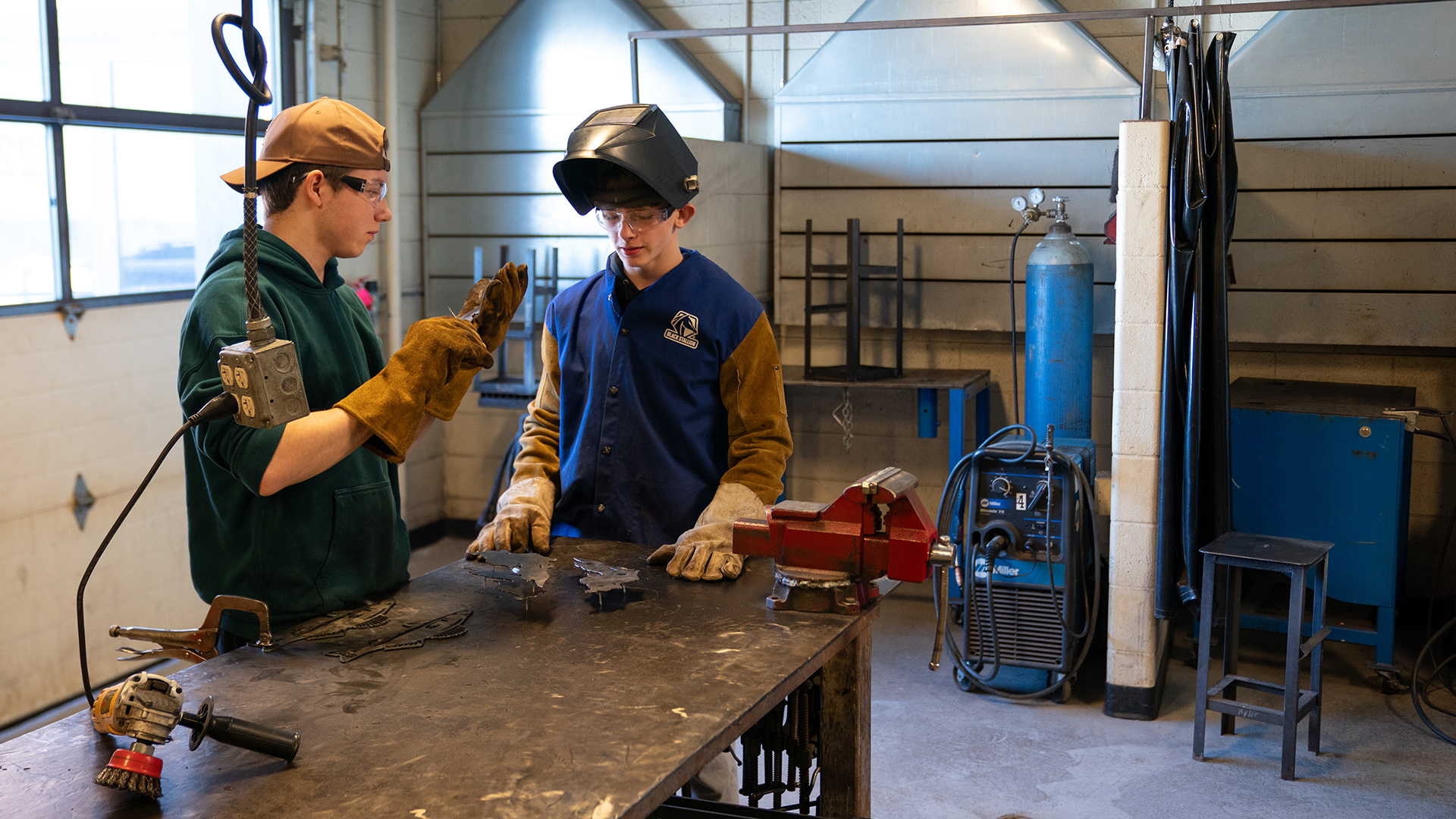 Two students, with one wearing welding gloves and another wearing a welding helmet that is flipped up and gloves stand on two sides of a work table with multiple tools on its surface, in a room with work benches, stools, a fuel tank attached to a machine with hoses and other equipment on a wall next to a garage door with windows.