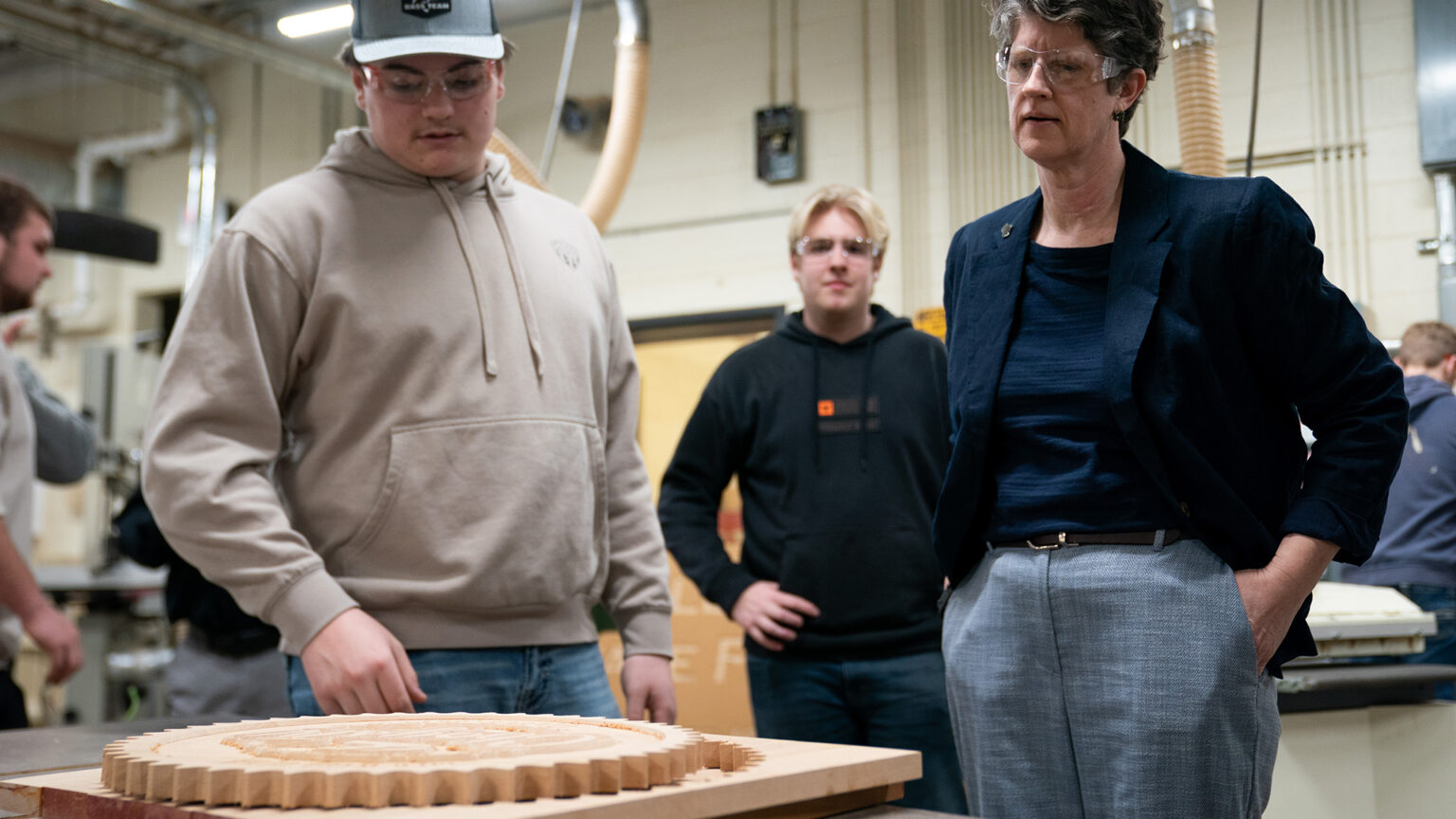 Thor Turra points toward a piece of carved wood in the shape of a gear on the surface of a table while standing next to Jill Underly, with other people standing in the background of a room with high-ceilings, exposed HVAC ducts and water pipes and overhead fluorescent lighting.
