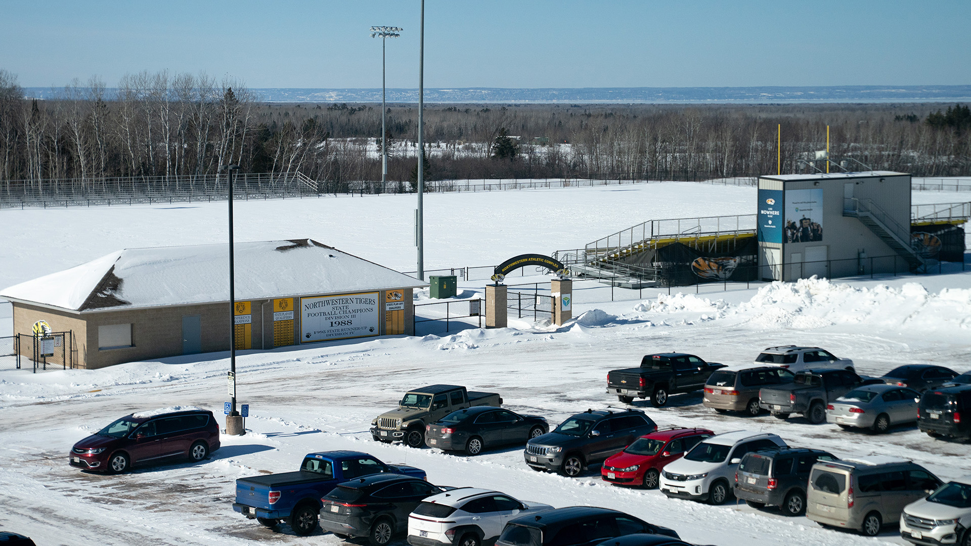 Multiple rows of vehicles are parked in a snow-covered parking lot in front of single-story building with a snow-covered roof next to a snow-covered field with stadium stands on either side and multiple light poles, with wooded areas in the background and a large body of water and bluffs on the horizon.