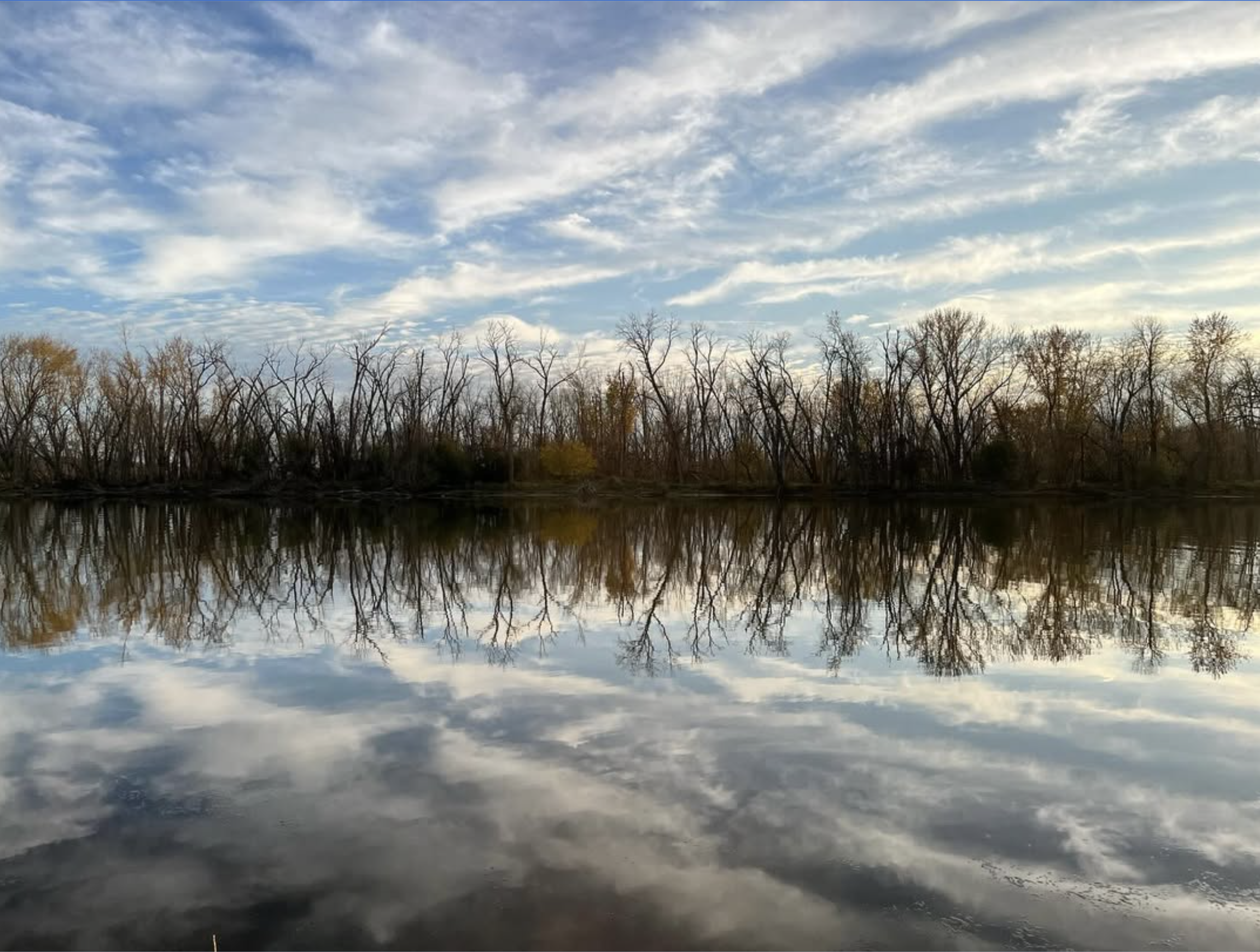 Calm Rock River on Lorine Niedecker's property with clear reflection of tall, leafless trees and a partly cloudy sky. The scene is serene, evoking a peaceful and tranquil atmosphere.