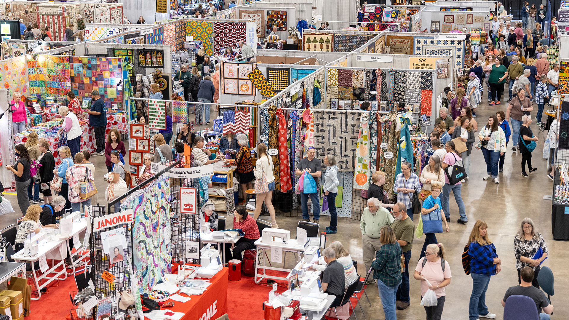 Overhead view of a packed quilt show exhibition hall, with hundreds of attendees browsing rows of colorful quilts displayed on grid walls and vendor booths, including a prominent Janome sewing machine display in the foreground.