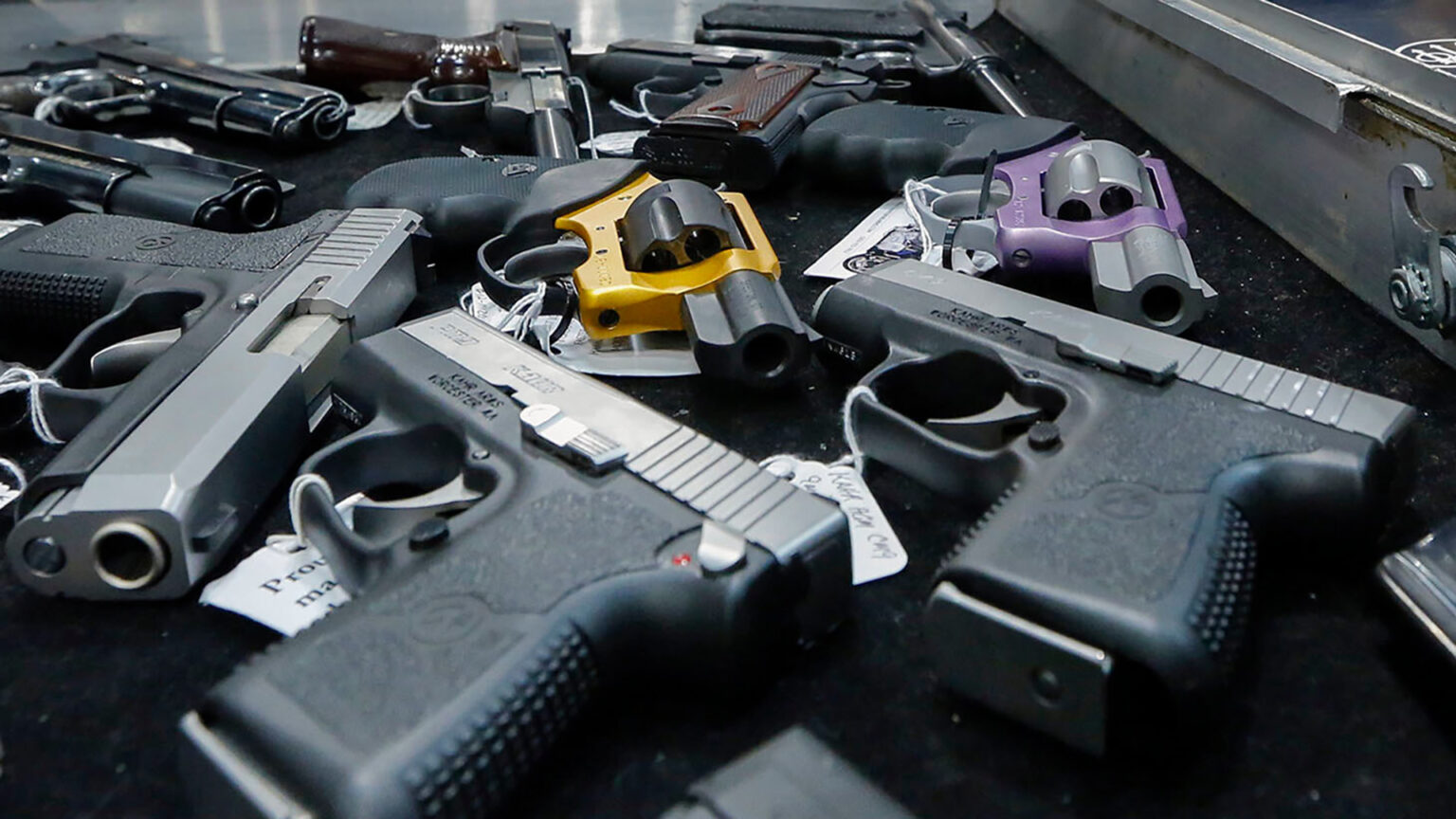 Multiple types of handguns with paper price tags attached to them sit on their sides on a table next to a metal case.