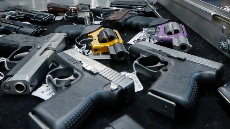Multiple types of handguns with paper price tags attached to them sit on their sides on a table next to a metal case.