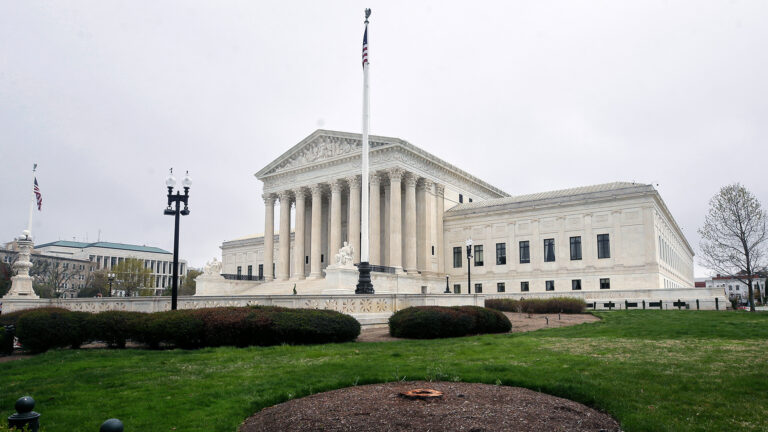 U.S. flags are hang on two flagpoles on either side of a plaza in front of a a multi-wing stone masonry building with steps leading up to an entrance framed by a portico of columns upholding a pediment with sculptures of figures, with trees on both sides in the background and a masonry wall behind a lawn with landscape plants and circular area covered by mulch in the foreground.