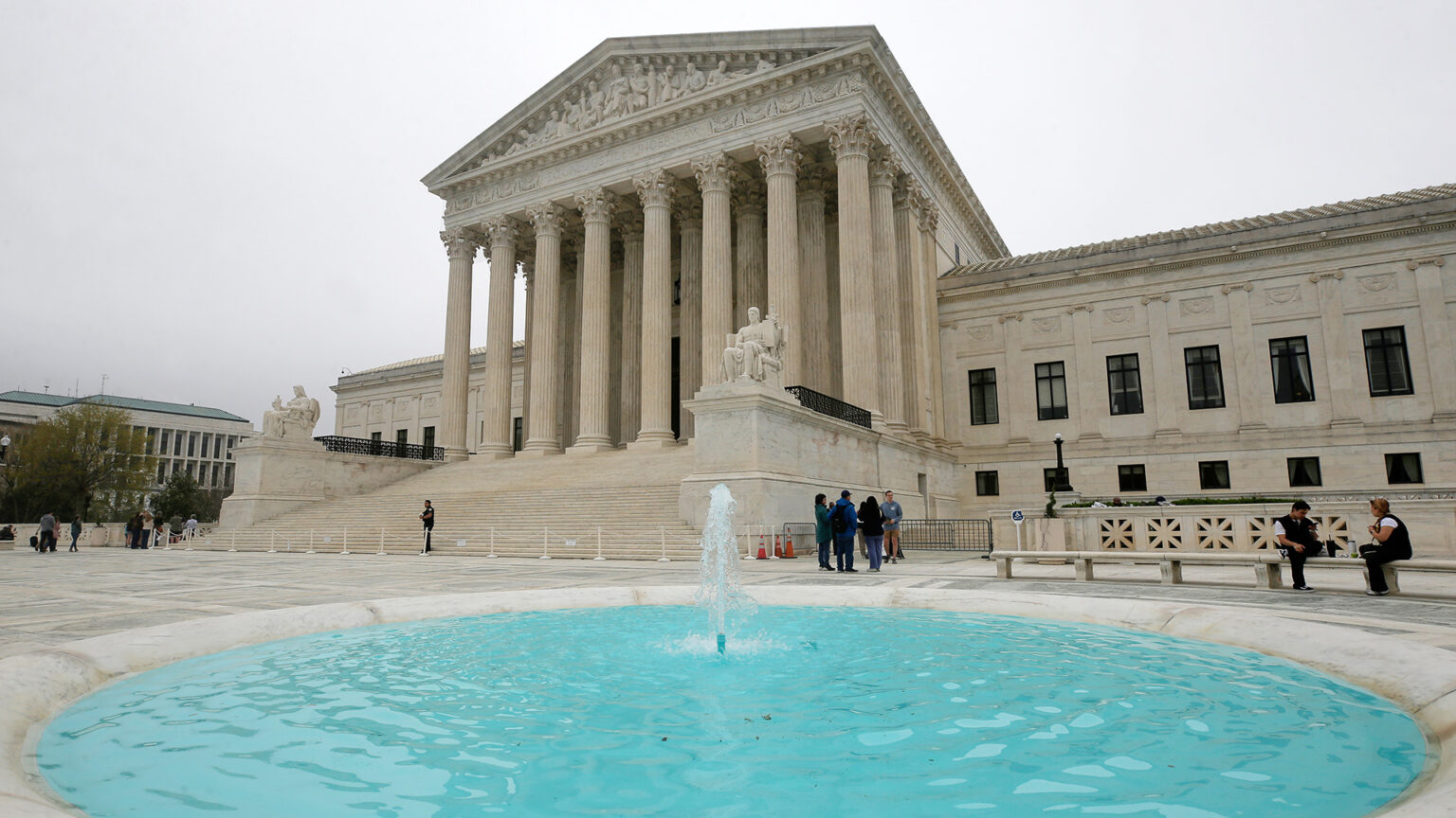 Multiple groups of people stand on a plaza and two people sit on benches in front of a multi-wing stone masonry building with steps leading up to an entrance framed by a portico of columns upholding a pediment with sculptures of figures, with a building and trees to the side in the background and a water fountain in the center of a shallow circular pool in the foreground.