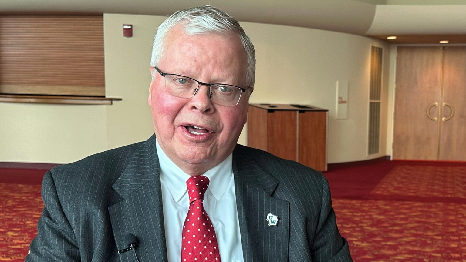 Jay Rothman speaks while sitting with a lavalier attached to one lapel and a Universities of Wisconsin logo pin attached to the other lapel of his suit coat, in a room with curved walls, a low-pile patterned carpet, and wood double doors, trash and recycling receptacle containers and rolling service window cover.