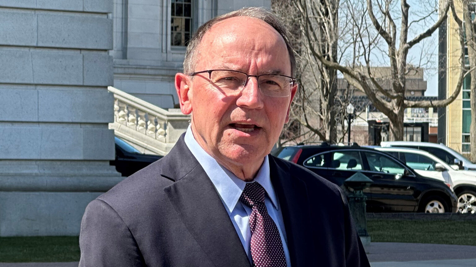 Tom Tiffany speaks while standing outside in front of a masonry building with a staircase with a balustrades, with parked vehicles, leafless trees and other buildings on one side in the background.