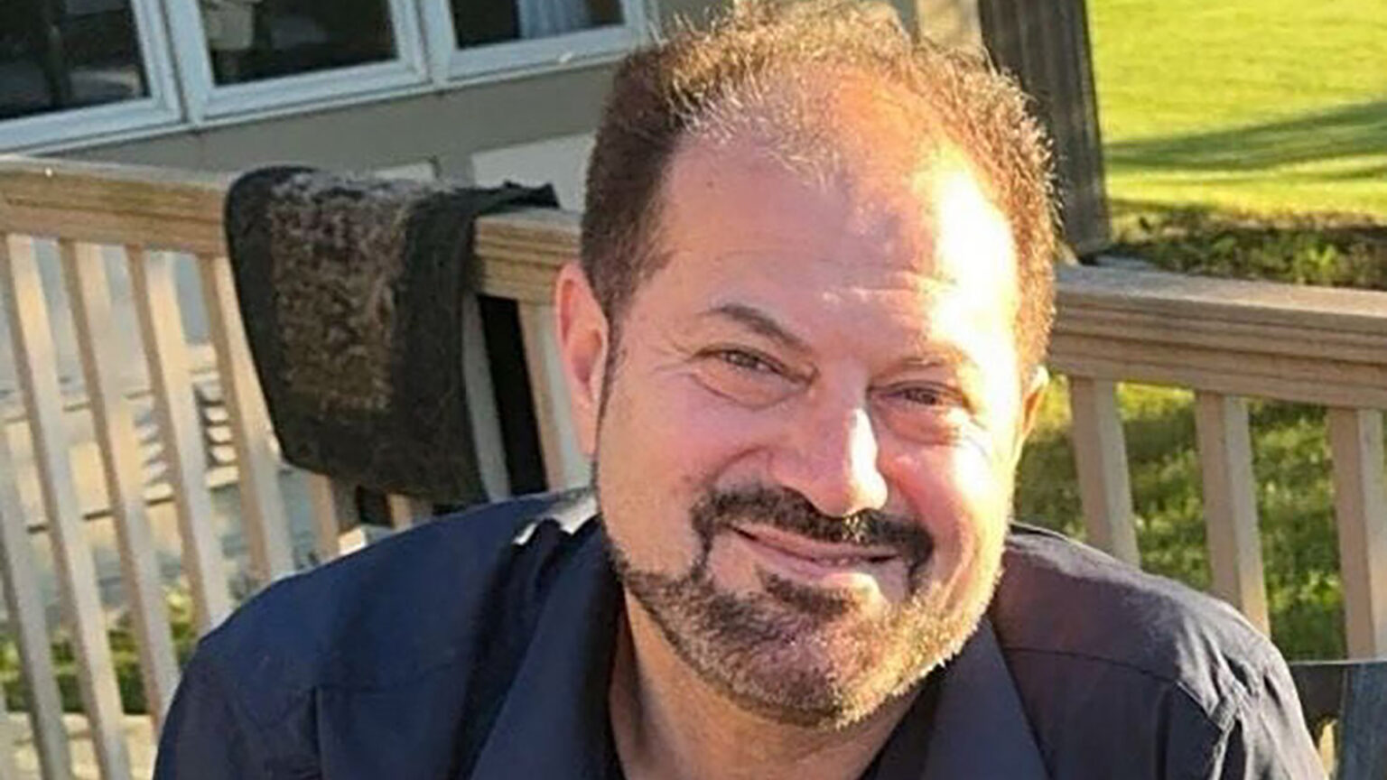 Salah Sarsour poses for a portrait with sunlight illuminating his face while sitting outside on a deck, with a wood railing, building with windows and lawn in the background.