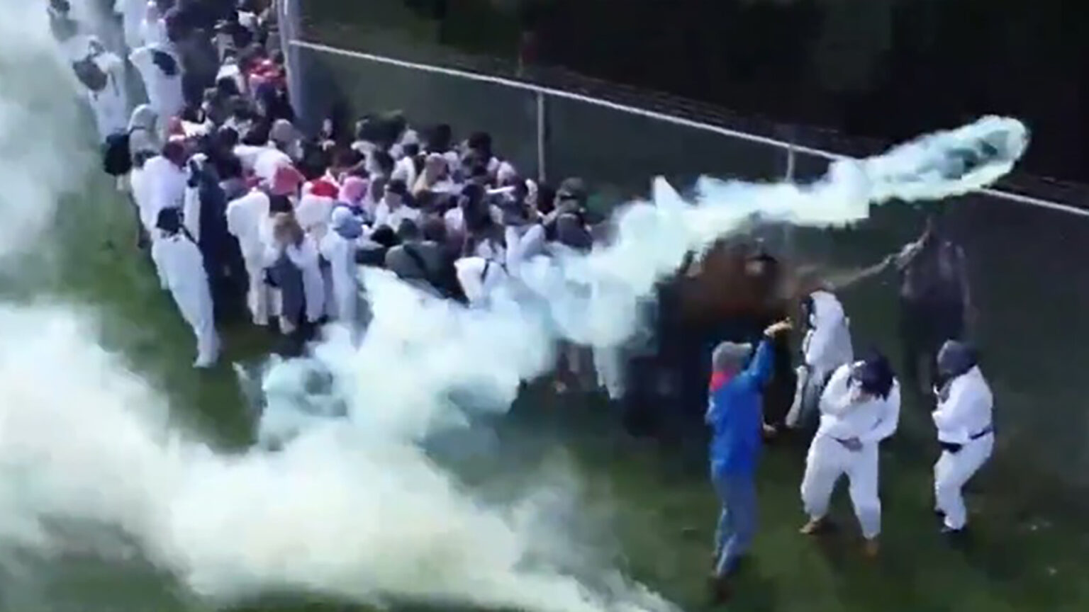 A group of people stand on one side of a chain-link fence topped by barbed wire, many wearing disposable coveralls with hoods, with one law enforcement officer standing on the other side of the fence using a can of pepper spray, and with clouds of tear gas streaming in the air, including one from an aloft canister.