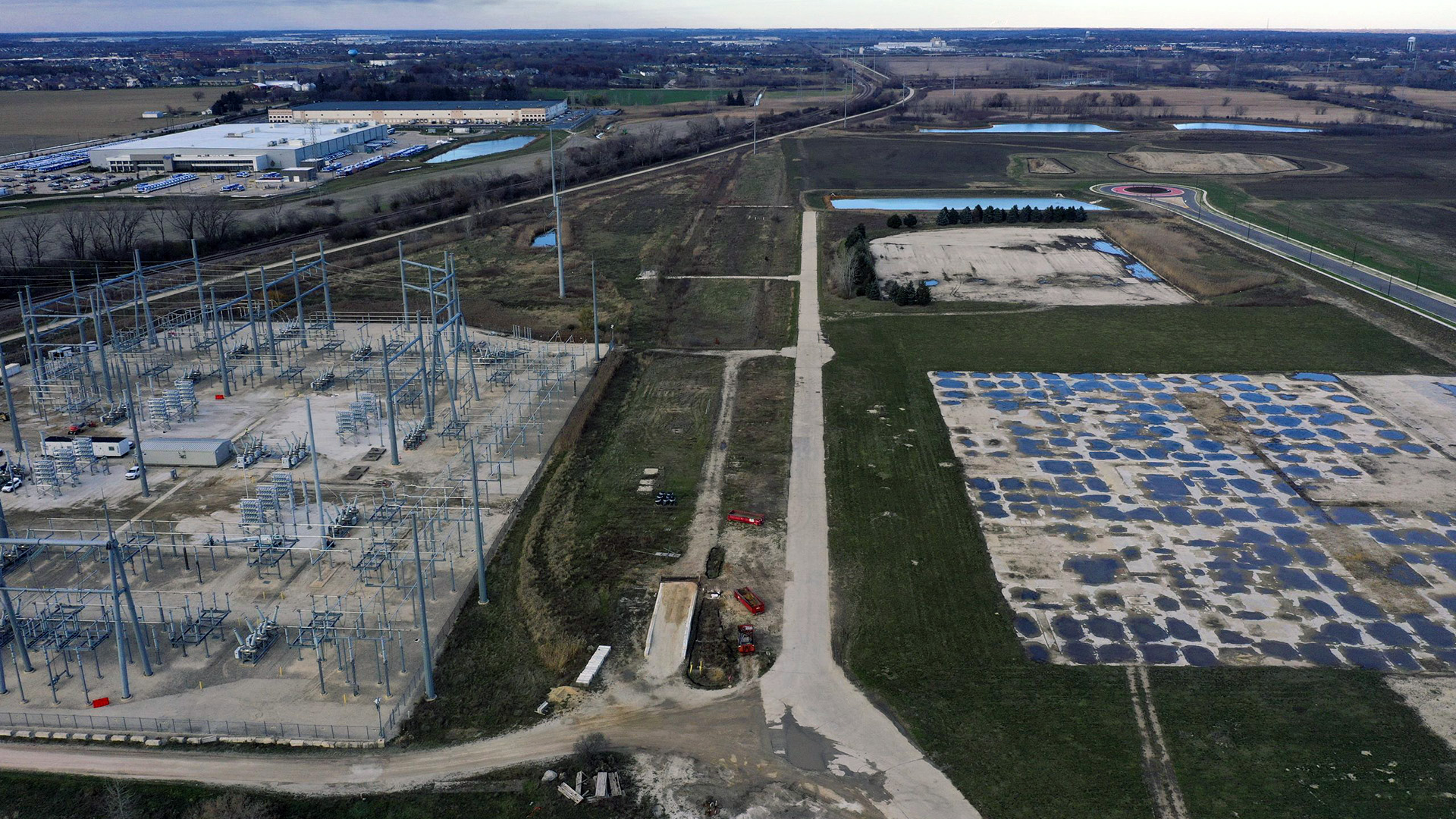 An aerial photo shows an electrical substation, a concrete pad marking the location of a former building, and multiple new buildings standing among fields, retention ponds and powerline towers, with fields and wooded areas in the background.