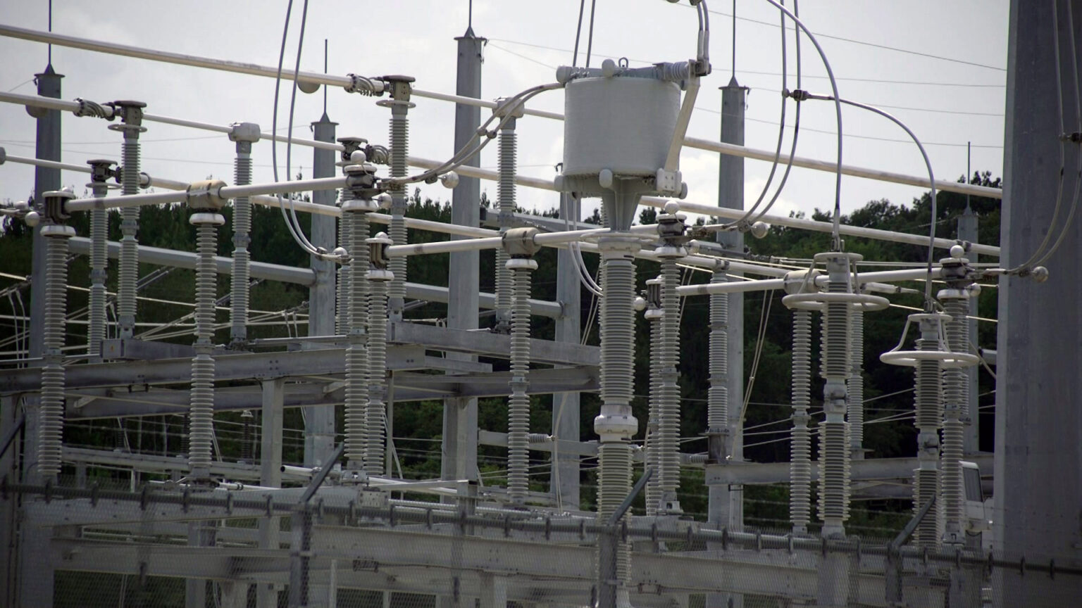 Multiple vertical towers and horizontal bars form part of an electrical substation, with a line of trees in the background.