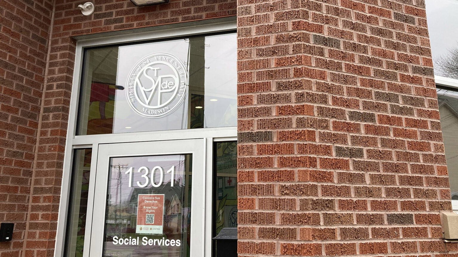 A graphic of the logo of Society of St. Vincent de Paul-Madison is affixed to a window above a metal-framed glass door with an address number, the word Social Services and a paper sign with a QR code affixed to its surface in a brick building with a window to the side, with reflections of adjacent buildings and utility poles visible in the door and windows.