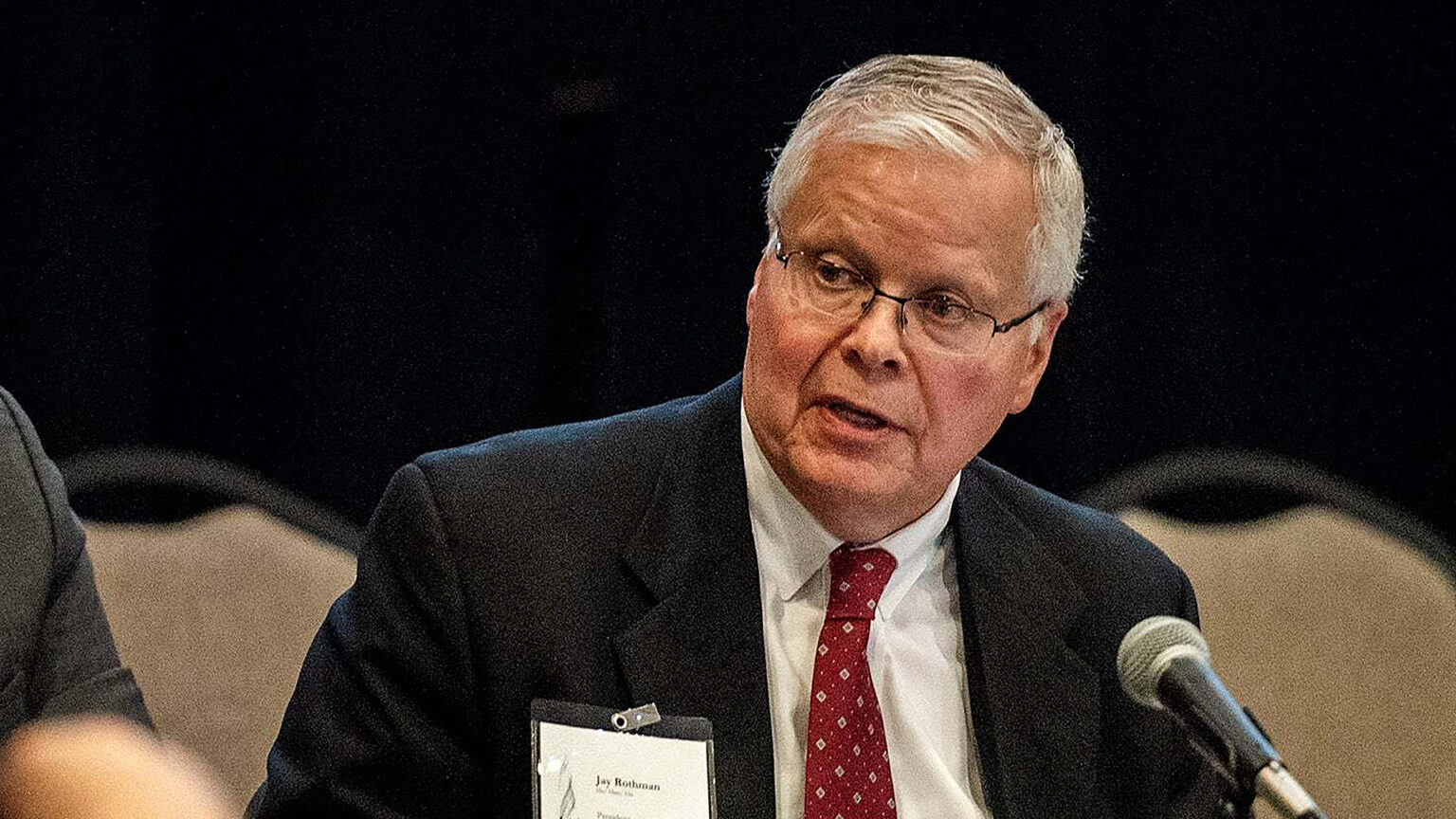 Jay Rothman sits and speaks into a mounted microphone, with an the arm of another person to his side and out-of-focus stackable chairs in the background.