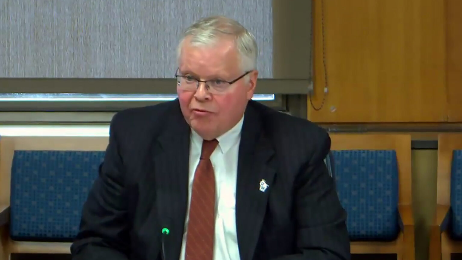 Jay Rothman sits and speaks into a microphone in a room with wood chairs with upholstered backs, wood-paneled walls and a closed window shade in the background.