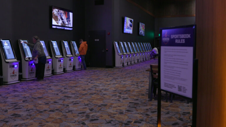 Two gamblers stand and one sits in front of two rows of self-service sports betting kiosks in a room with multiple flatscreen televisions mounted to walls, a low-pile carpet and an out-of-focus sign with the title text Sportsbook Rules and other writing in the foreground.