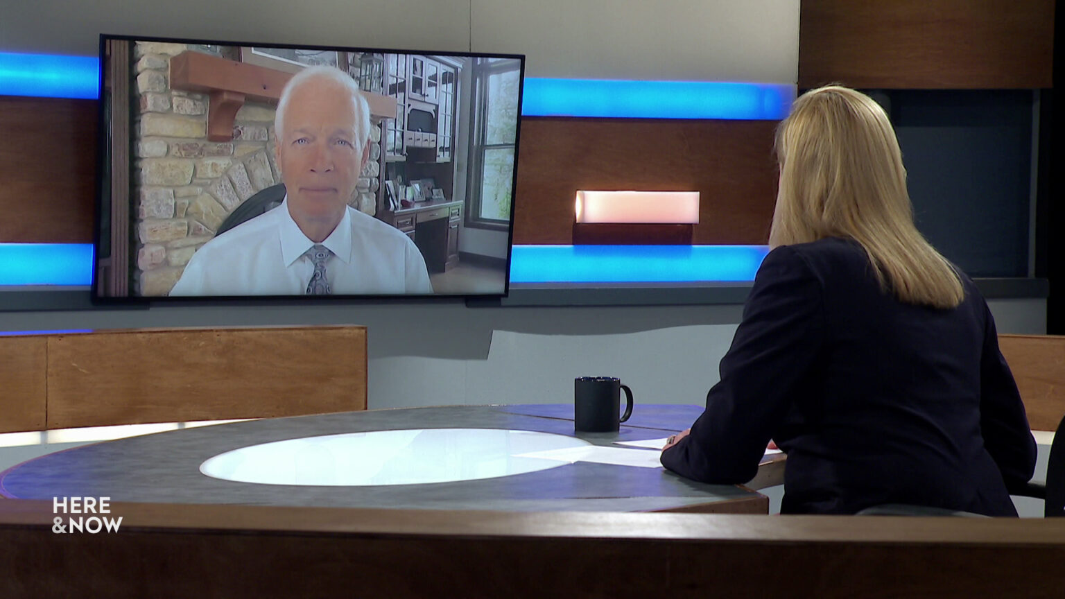 Frederica Freyberg sits at a desk on the Here & Now set and faces a video monitor showing an image of Ron Johnson.