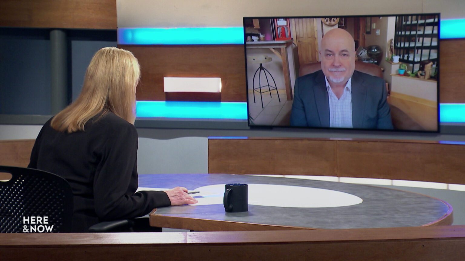 Frederica Freyberg sits at a desk on the Here & Now set and faces a video monitor showing an image of Mark Pocan.