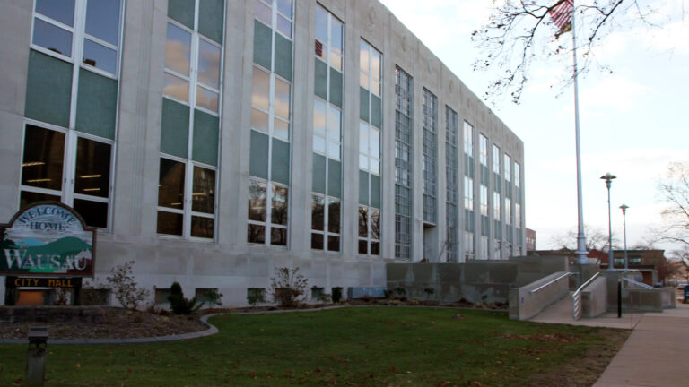 A weathered wood sign with stylized images of hills and trees and the words Welcome Home to Wausau and City Hall stands in a mulched garden bed in front of a multi-story masonry building with large glass windows in front of small grass lawn, a concrete ramp and a flagpole with a U.S. flag, withe light poles, buildings and leafless trees in the background to the side.