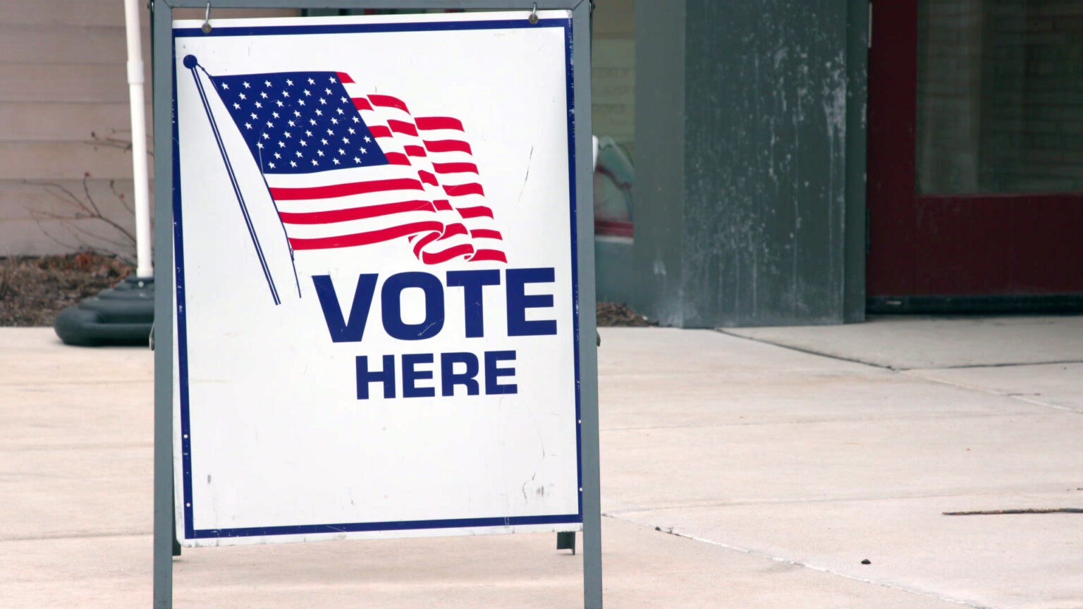 A sandwich board sign with an illustration of a U.S. flag and the words Vote Here stands on a concrete sidewalk in front of a building.
