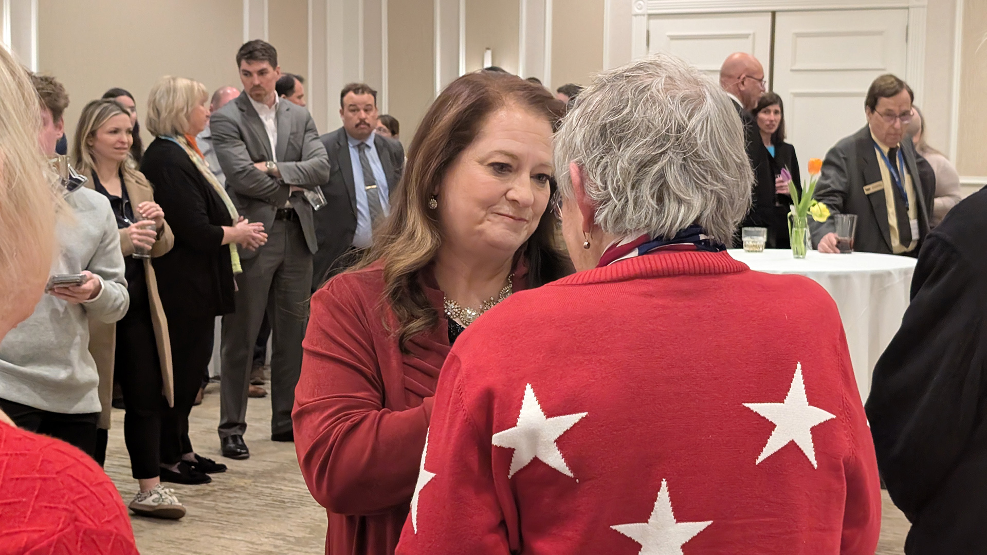 Maria Lazar speaks while standing and facing another person in the foreground, with other people standing next to each other and around a table in the background in a room with closed double doors.