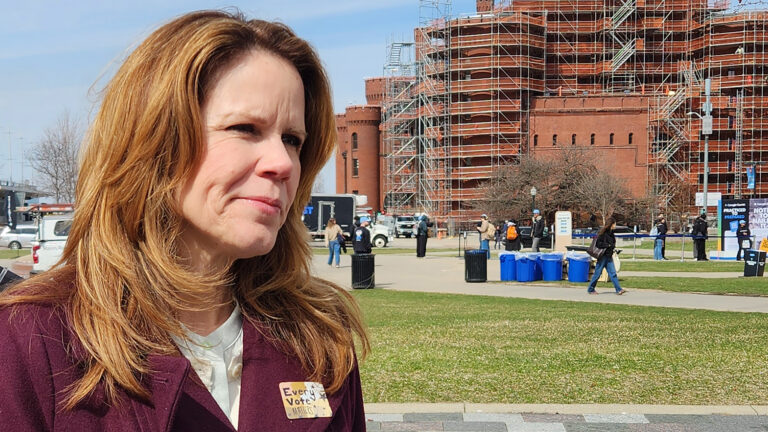 Chris Taylor speaks and wears a sticker with the words Every Vote Matters while standing outside, with a lawn and walkways, trash receptacles, parked vehicles and a Romanesque Revival building surrounded by construction scaffolding in the background. Chris Taylor speaks and wears a sticker with the words Every Vote Matters while standing outside, with a lawn and walkways, trash receptacles, parked vehicles and a Romanesque Revival building surrounded by construction scaffolding in the background.