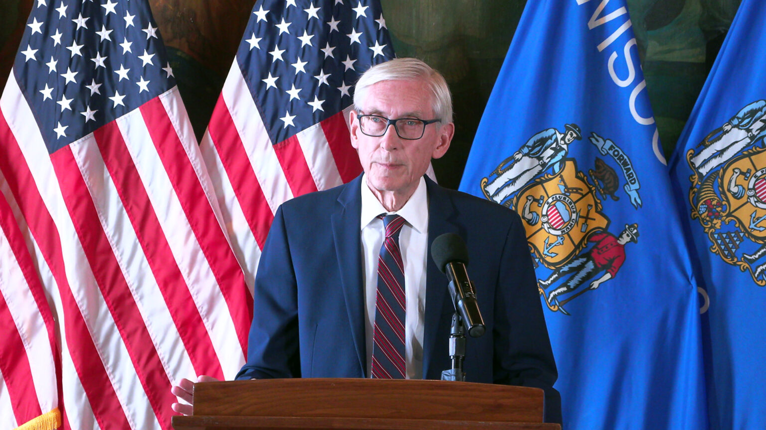 Tony Evers speaks into a microphone mounted to the top of a wood podium while standing in front of a row of U.S. and Wisconsin flags in front of a large painting. Tony Evers speaks into a microphone mounted to the top of a wood podium while standing in front of a row of U.S. and Wisconsin flags in front of a large painting.