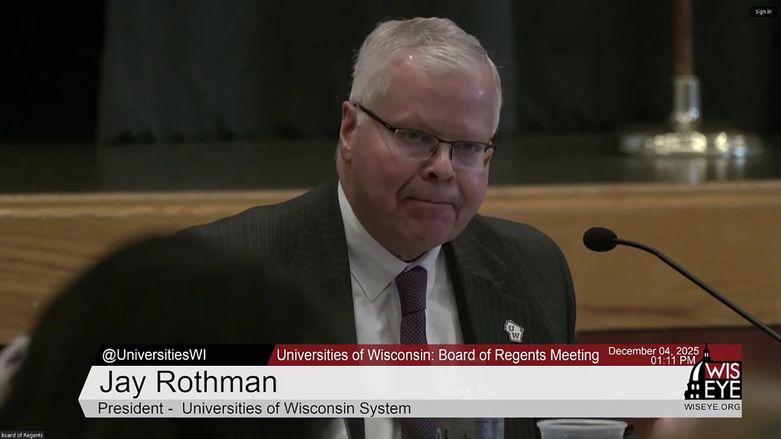 Jay Rothman speaks while seated and facing a mounted microphone, in a room with an out-of-focus wood shelf with the base of a light fixture on its surface, with a video graphic at bottom including the text Jay Rothman and President - Universities of Wisconsin System.