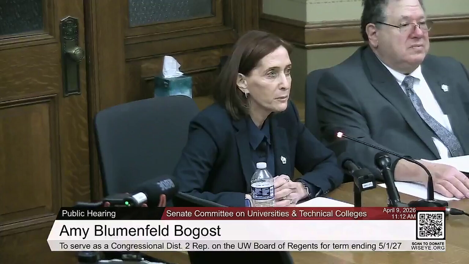 Amy Blumenfeld Bogost speaks while seated at a table and facing multiple mounted microphones and a video camera with a shotgun microphone, with Timothy Nixon seated to her side, in a room with wood trim on the wall and wood double doors with a brass doorknob and glazed glass windows, with a video graphic at bottom including the text Senate Committee on Universities & Technical Colleges, Amy Blumenfeld Bogost and To serve as a Congressional Dist. 2 Rep. on the UW Board of Regents for term ending 5/1/27.