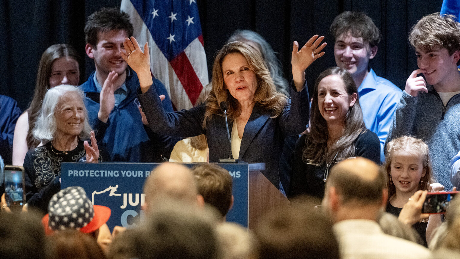 Chris Taylor waves with both hands while standing behind a podium with a microphone affixed to its top, while other people stand behind her and facing her with a U.S. flag in the background.
