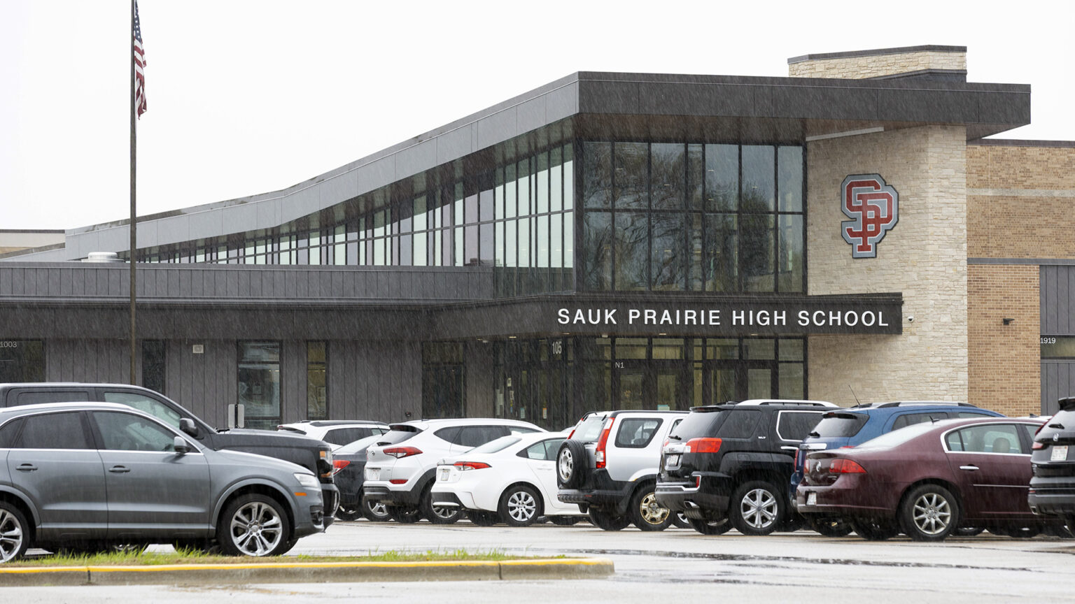 Rain falls on multiple vehicles parked in rows in an asphalt-covered parking lot in front of a multi-story brick and glass building with the a letter sign reading Sauk Prairie High School on  an awning below a logo consisting of an overlapping S and P, with a U.S. flag on a flagpole to the side.