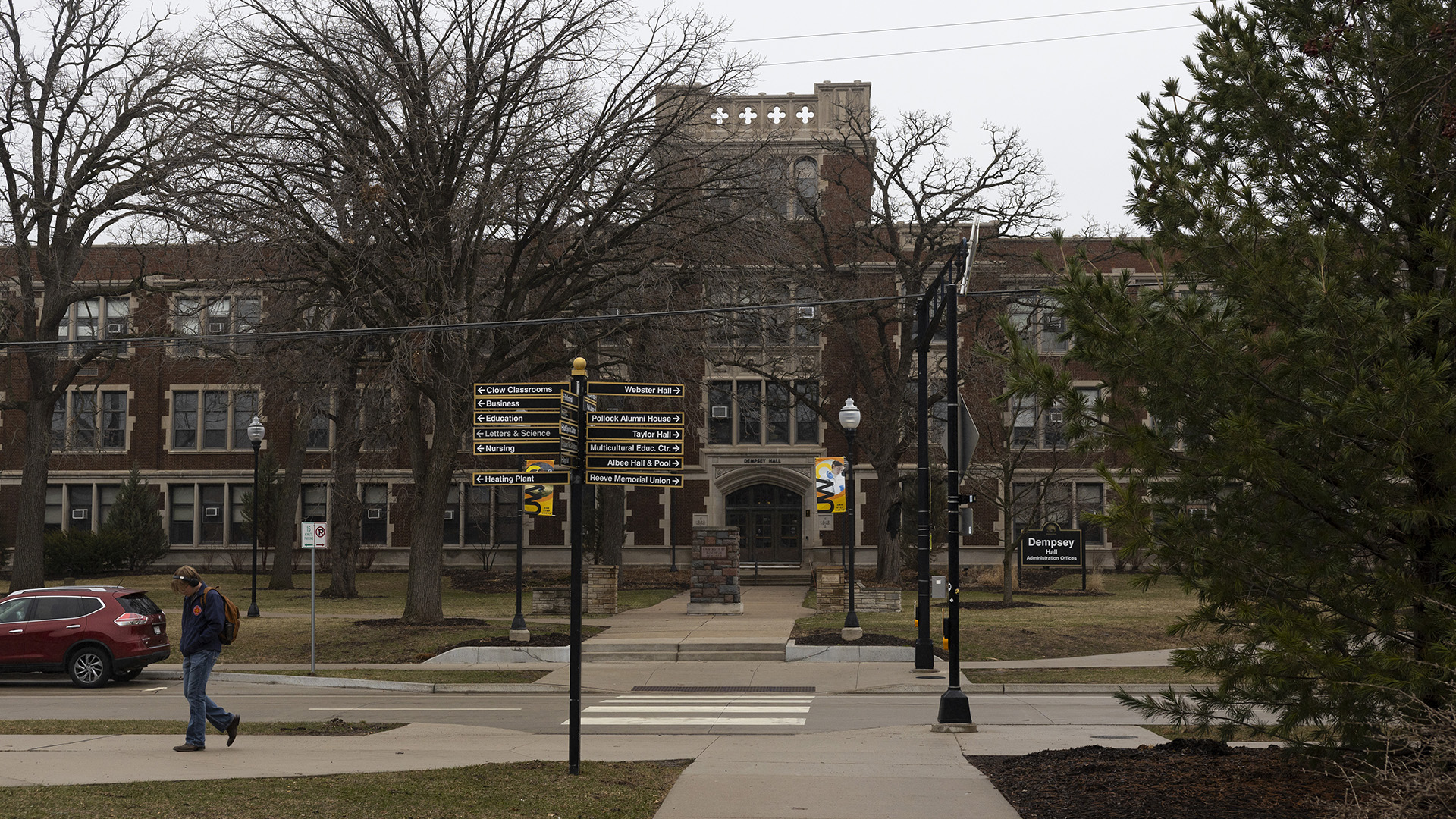 A pedestrian walks on a sidewalk away from a crosswalk over a street with a vehicle parked on one side in front of a lawn with leafless trees and a multi-story masonry and brick building with two wings and a central tower in the background, with traffic signal poles and sign poles on either side of the street and a coniferous tree to one side in the foreground.
