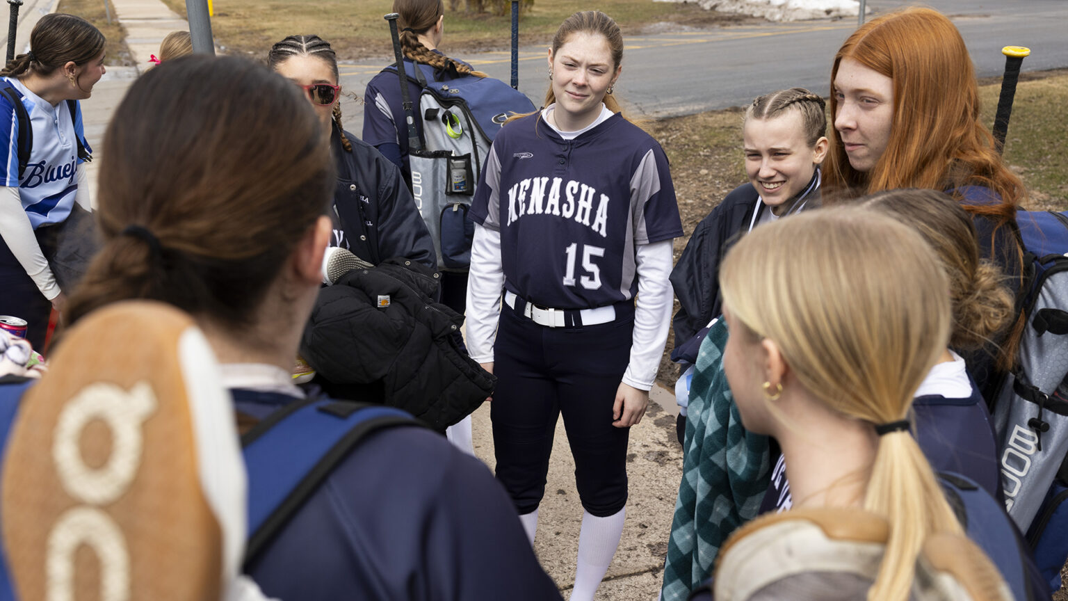 Teagan Massey-Plamann, wearing a baseball uniform with the word Menasha and numeral 15 on the front of the jersey, stands on a sidewalk among other people, several with backpacks with stowed softball bats, with lawns, a road and sidewalk in the background.
