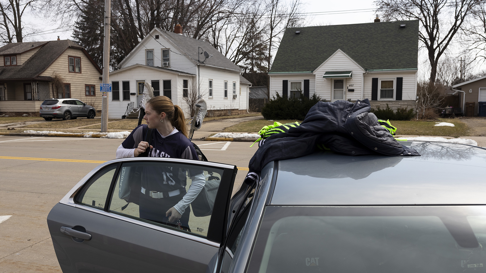 Teagan Massey-Plamann, wearing a baseball uniform with the word "Menasha" and numeral "15" on the front of the jersey, and carrying bags with cleats attached to the handles of two upside-down stowed softball bats, stands behind an open rear passenger side door of a sedan with a jacket and other items on its roof, with a street and row of different two-story houses with detached garages, front lawns and leafless trees in the background.