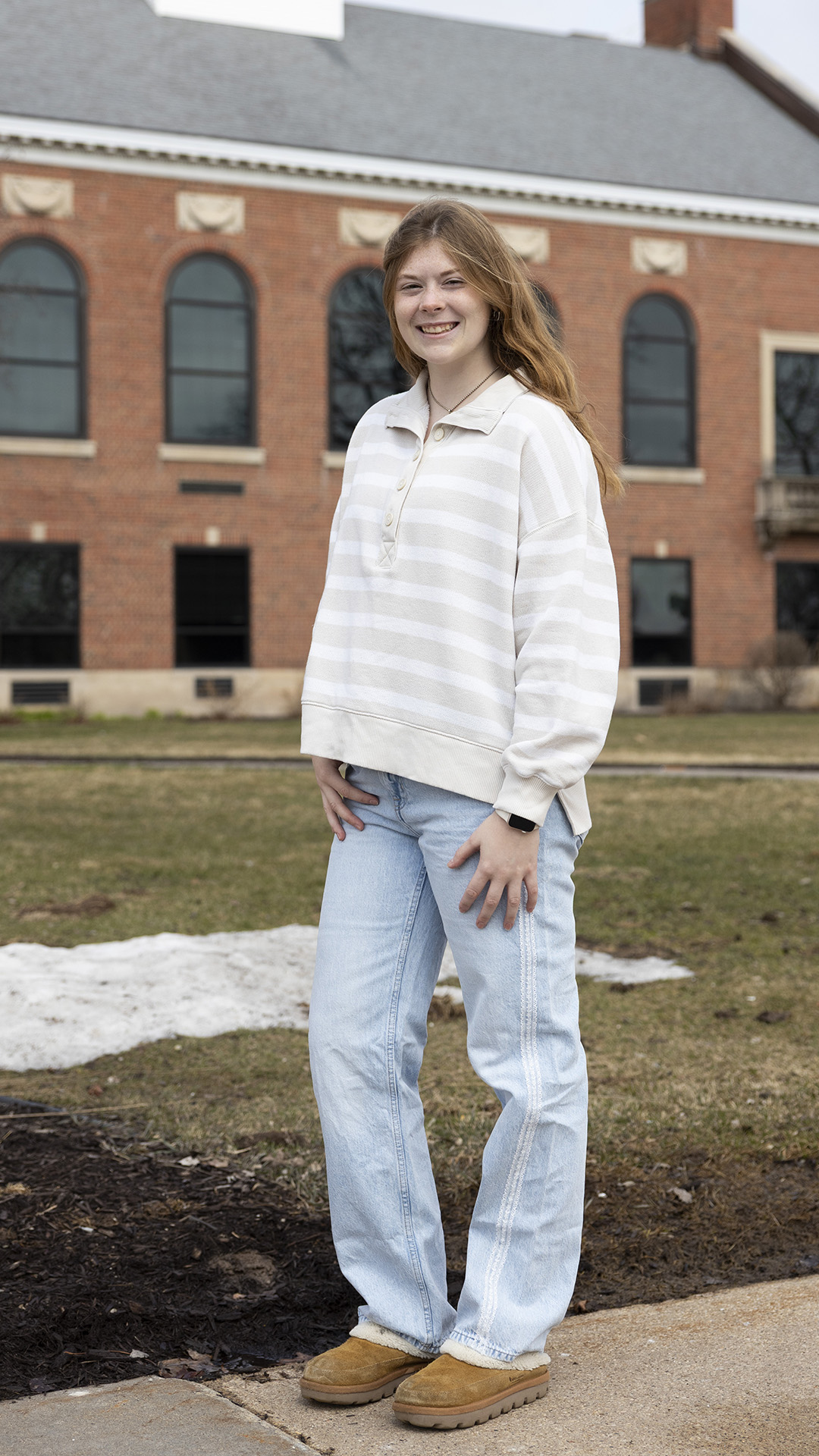 Teagan Massey-Plamann poses for a portrait while standing on a sidewalk in front of a lawn with a small snowbank in front of a multi-story brick building with arched windows on its second level and a pitched roof.
