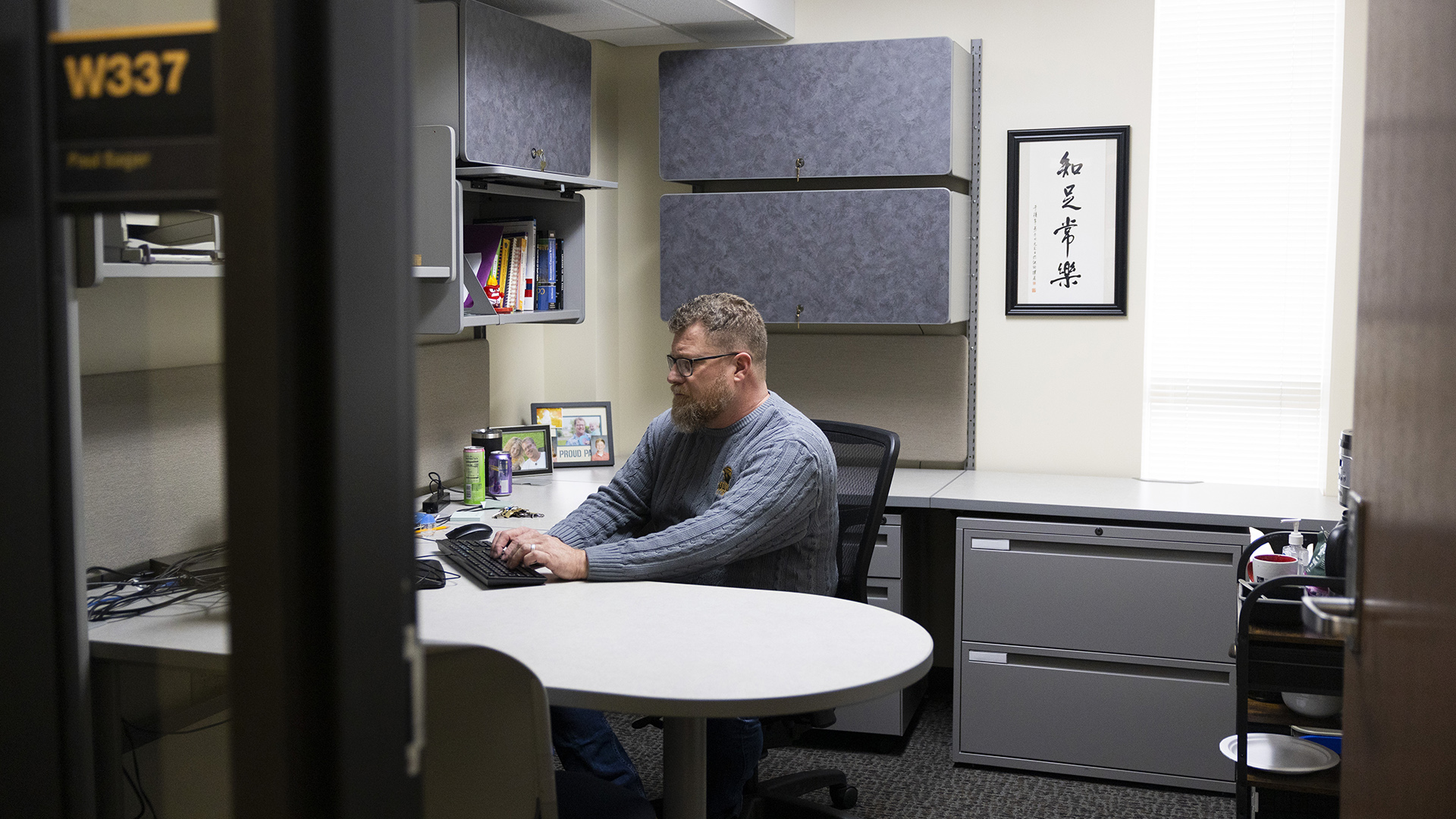 Paul Sager sits in a rolling desk chair and types on a keyboard on a desk surface, with additional sections of the desk behind him above metal file cabinets and with additional cabinets mounted to two walls next to a framed work of calligraphy and a window, with an out-of-focus metal and glass doorframe with a room label reading "W337" and an open wood door with a metal passage lever in the foreground.