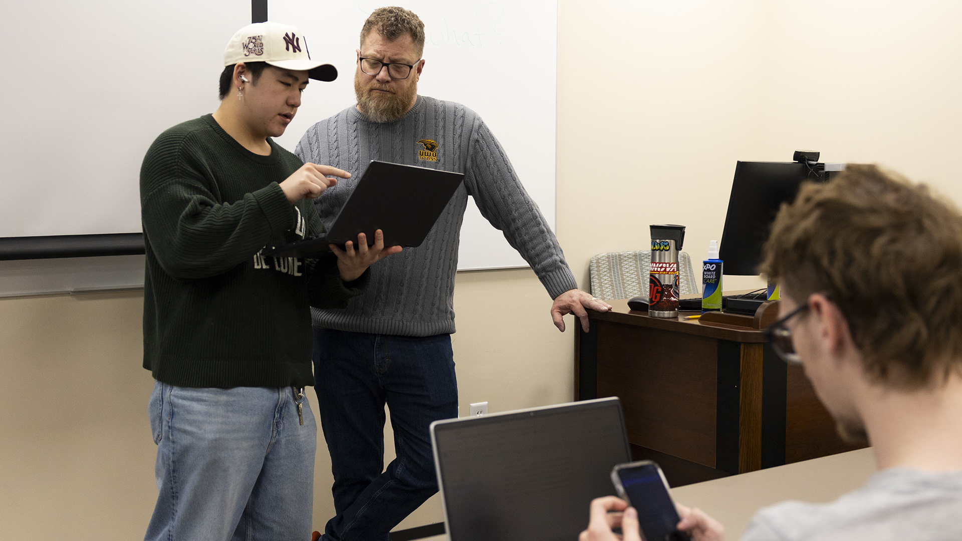 Hugh Thao holds a laptop computer in his left hand and points to its screen with his right hand while standing next to Paul Sager, whose left hand is on the corner of a wood desk with a beverage container and whiteboard cleaner bottle on its surface next to a computer monitor, with a whiteboard on the wall behind them and an out-of-focus person holding a smart phone in both hands while seated in front of a laptop computer in the foreground.