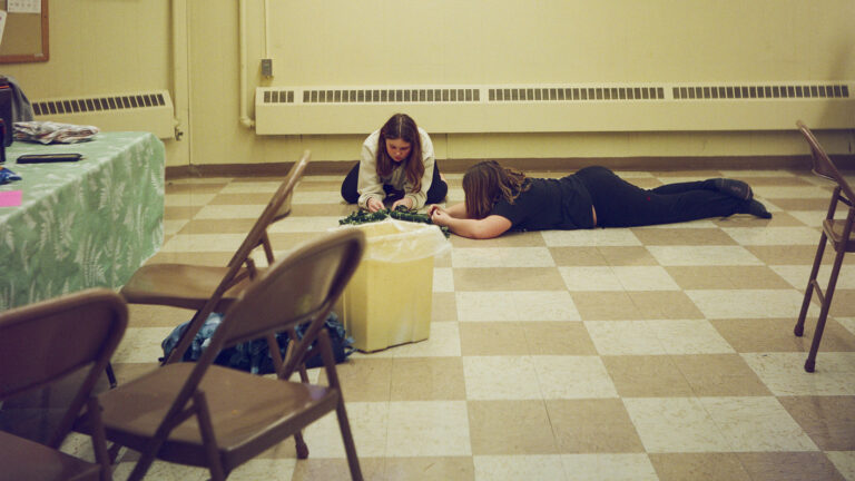 One person crouches and another lays face-down on vinyl tile flooring, with a table covered by a tablecloth and items on its surface, folding metal chairs and a plastic trash container in the foreground, and radiators along walls in the background. One person crouches and another lays face-down on vinyl tile flooring, with a table covered by a tablecloth and items on its surface, folding metal chairs and a plastic trash container in the foreground, and radiators along walls in the background.
