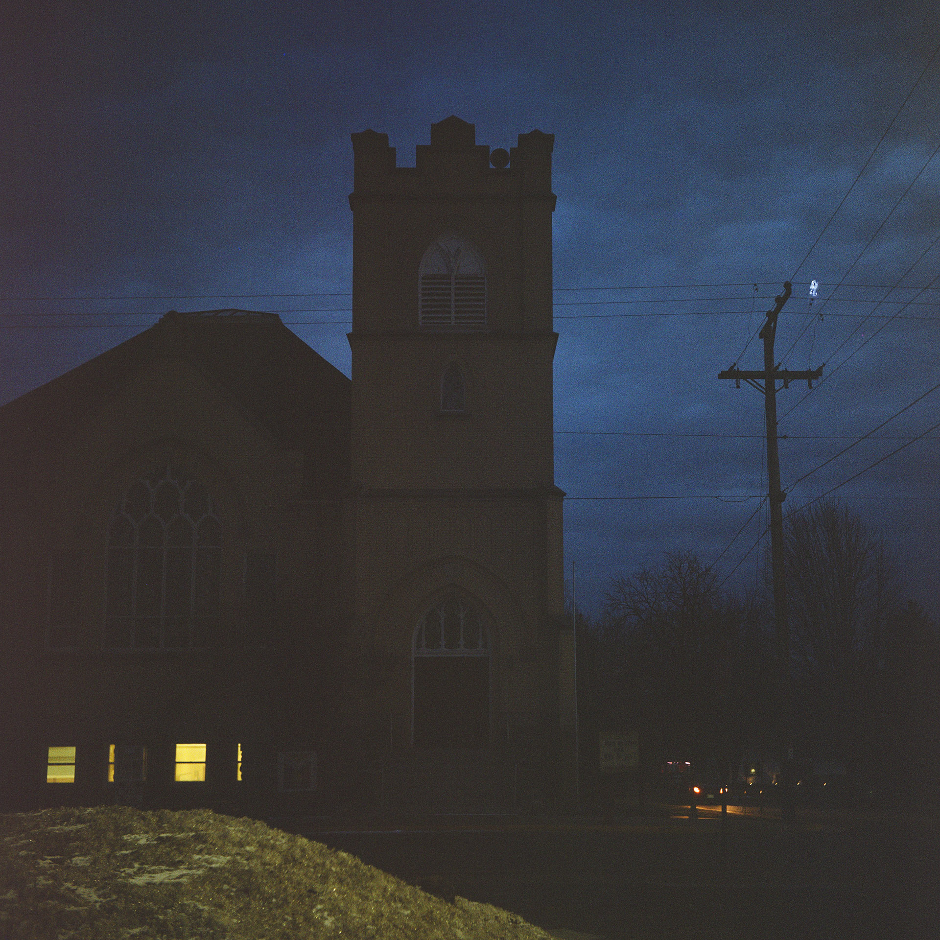 Light shines in two lower windows of a multistory church with a large arched window of a nave next to a three-level crenellated tower, with power poles and lines silhouetted against a dark and cloudy sky.