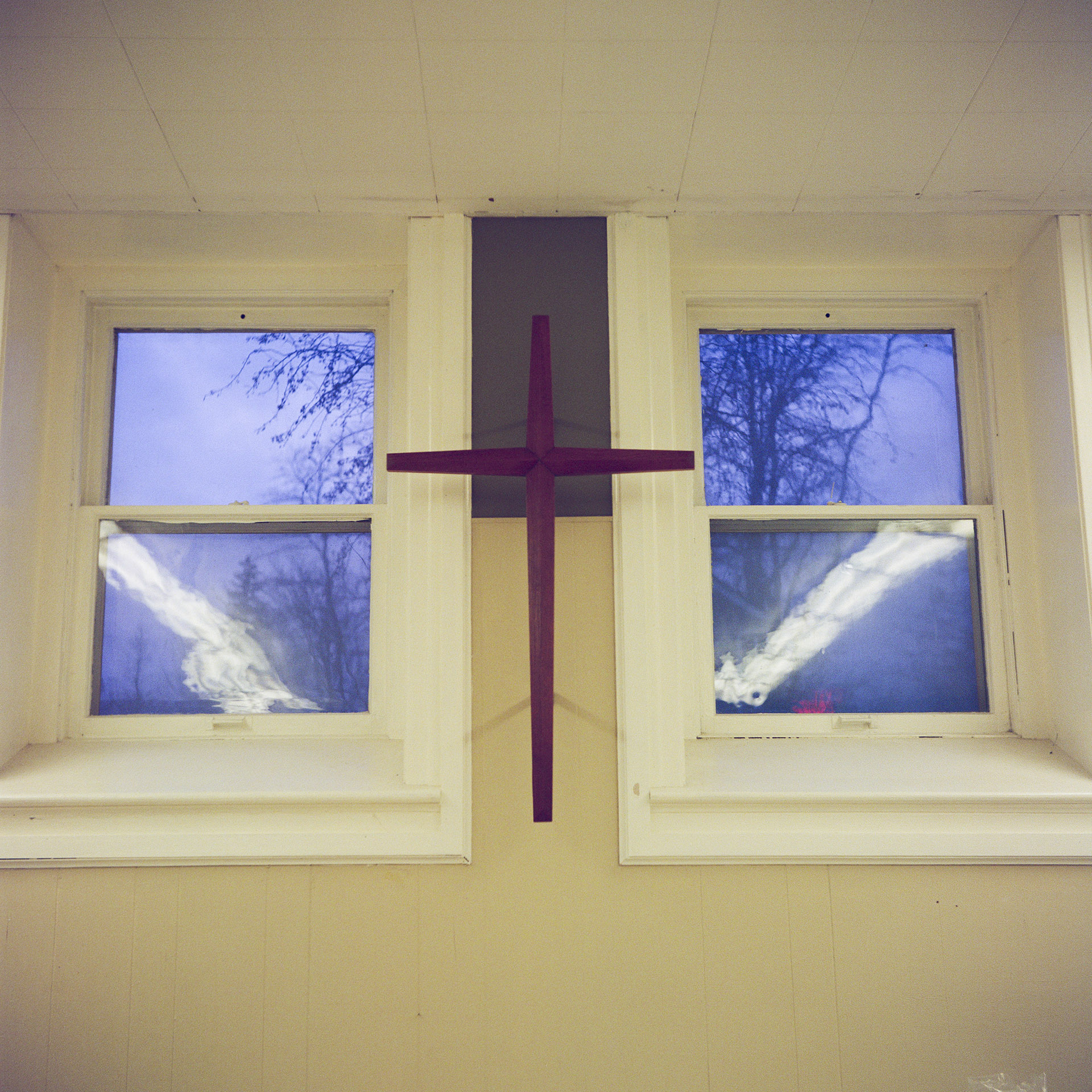 A cross is mounted on a wall between two closed windows with leafless trees visible outside.